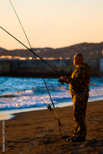 Elderly man fishes with fishing rod on ocean shore at sunset. Fisherman holds fishing rod in his hands and catches fish by sea. Leisure of pensioner. Hobby: fishing.