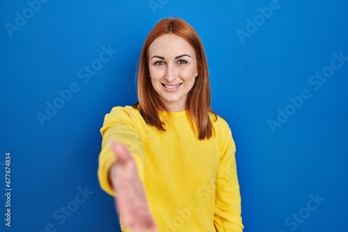Young woman standing over blue background smiling friendly offering handshake as greeting and welcoming. successful business.