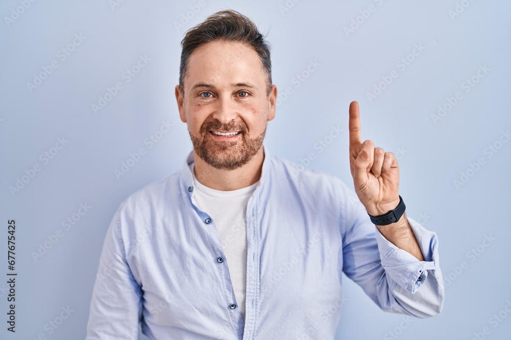 Middle age caucasian man standing over blue background showing and pointing up with finger number one while smiling confident and happy.