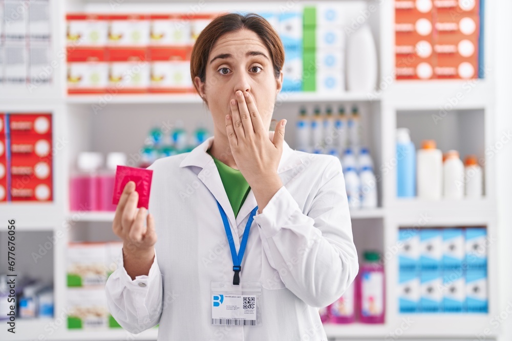 Brunette woman working at pharmacy drugstore holding condom covering mouth with hand, shocked and afraid for mistake. surprised expression