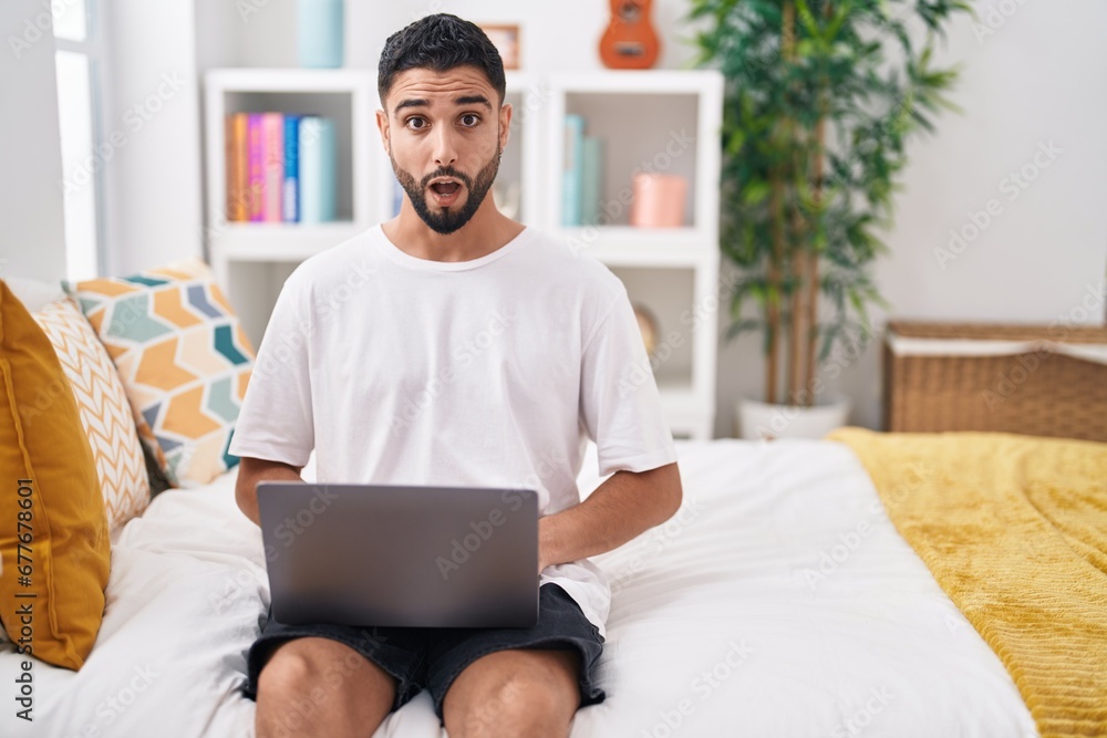 Hispanic young man using computer laptop sitting on the bed scared and amazed with open mouth for surprise, disbelief face Hispanic young man using computer laptop sitting on the bed scared and amazed with open mouth for surprise, disbelief face
