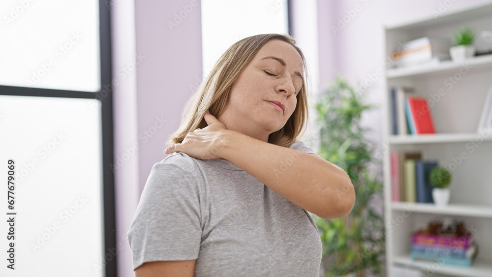 Portrait of a distressed, attractive, young blonde woman suffering from a severe neck ache, standing unhappily indoors within her cozy home Portrait of a distressed, attractive, young blonde woman suffering from a severe neck ache, standing unhappily indoors within her cozy home