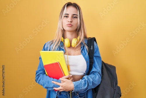 Young blonde woman wearing student backpack and holding books looking at the camera blowing a kiss on air being lovely and sexy. love expression.