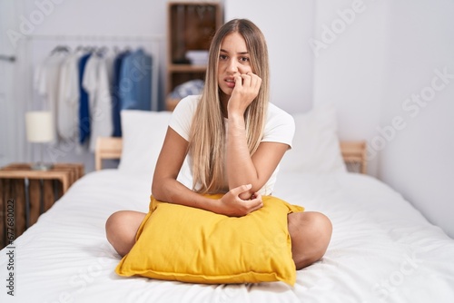 Young blonde woman sitting on the bed with pillow at home looking stressed and nervous with hands on mouth biting nails. anxiety problem.