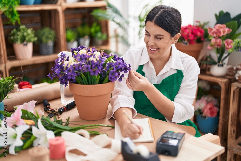 Young beautiful hispanic woman florist writing on notebook touching plant at flower shop