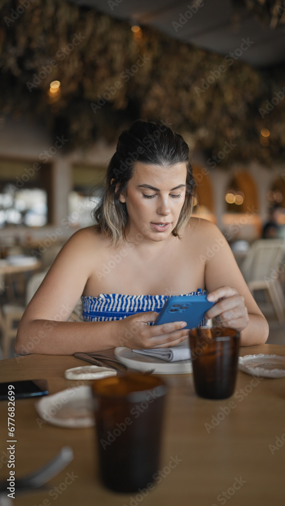 Young hispanic woman using smartphone sitting on the table at the restaurant