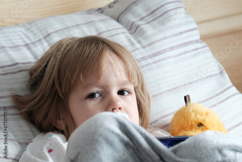 A small fair-haired, European-looking girl of two years old lies in bed with a pacifier, looks, plays on the phone. Concept of gadgets and their impact on children's health.