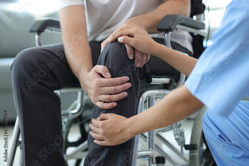 Female doctor diagnosing knee pain of male patient in wheelchair in hospital examination room.