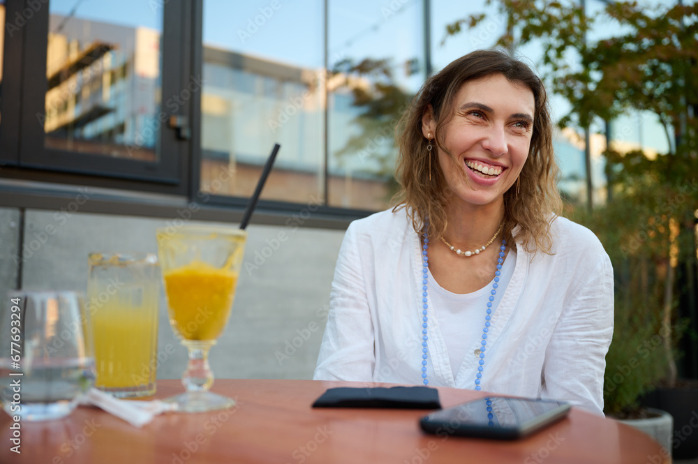 Young woman in a white shirt and a necklace around her neck on a bright sunny day in a cafe with greenery in the background is drinking fresh juice and actively talking about something