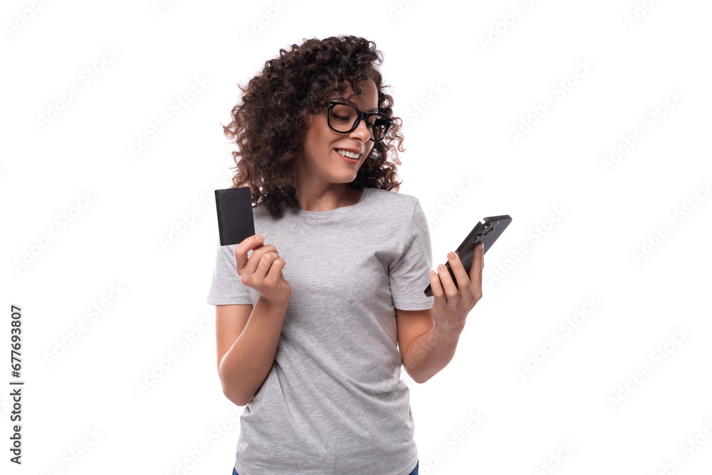 cute slim curly woman with black hair dressed in casual gray t-shirt holding smartphone and plastic credit card cute slim curly woman with black hair dressed in casual gray t-shirt holding smartphone and plastic credit card