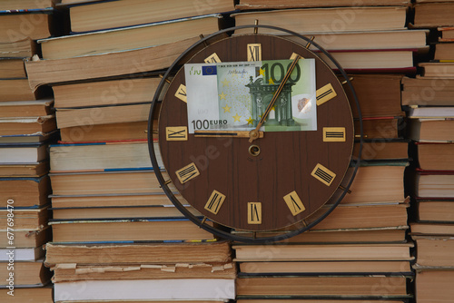 Large clock with Roman numerals on the clock face against a backdrop of stacked books, and one hundred euros on the clock face. Economic education, paid education, high tuition fees