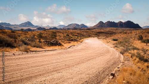 View of the Namib Desert in northern Erongo  Namibia  with mountains in the background and a car driving on a  gravel road  kicking up dust behind it.
