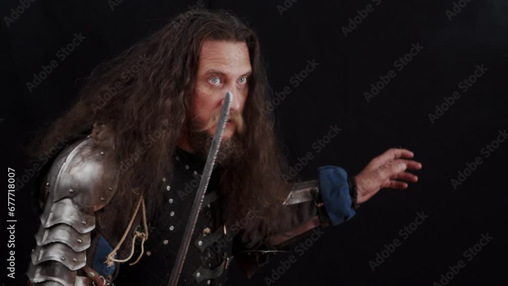 A charismatic man in the armor of a medieval knight beats with a sword on a black background. Slow motion. A charismatic man in the armor of a medieval knight beats with a sword on a black background. Slow motion.