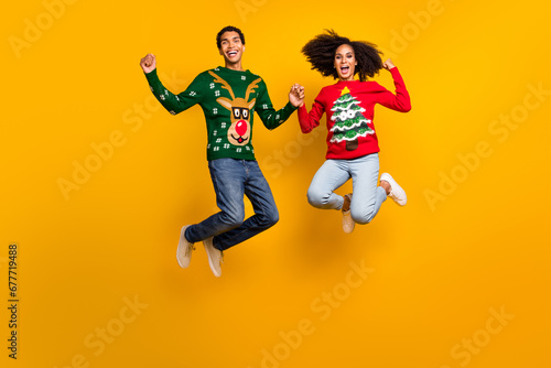 Full body photo of two delighted american people jumping hold arms raise fists x-mas sale isolated on yellow color background