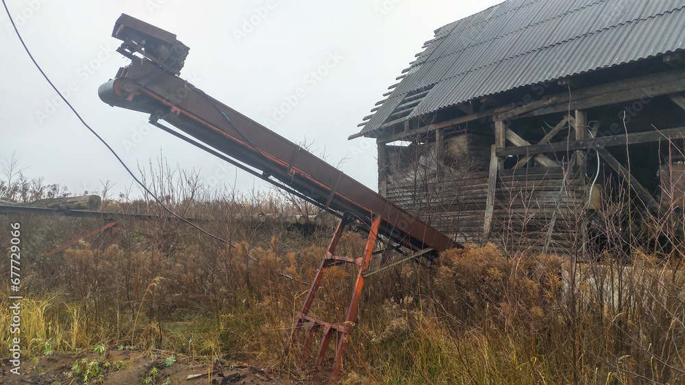 An abandoned board shed with a slate roof stands amidst a thicket of dry grass and bushes. A belt conveyor with an electric motor is mounted on a metal stand in front of it Cloudy weather and grey sky An abandoned board shed with a slate roof stands amidst a thicket of dry grass and bushes. A belt conveyor with an electric motor is mounted on a metal stand in front of it Cloudy weather and grey sky