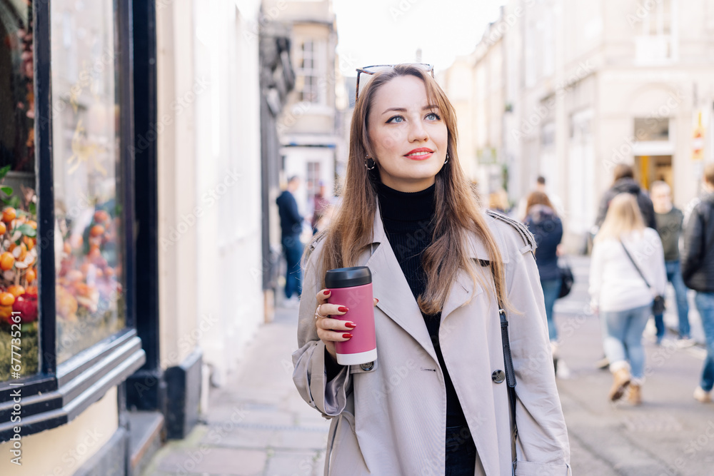 Young smiling woman in trench coat enjoying hot coffee drink in reusable cup while walking on the city streets decorated for the autumn holidays. Happy fall cozy mood, seasonal fashion style trend. Young smiling woman in trench coat enjoying hot coffee drink in reusable cup while walking on the city streets decorated for the autumn holidays. Happy fall cozy mood, seasonal fashion style trend.