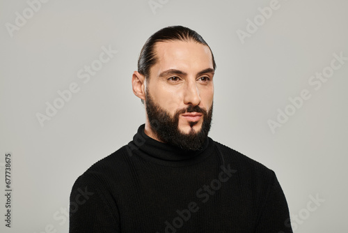 portrait of good looking arabic man with beard posing in black turtleneck on grey background