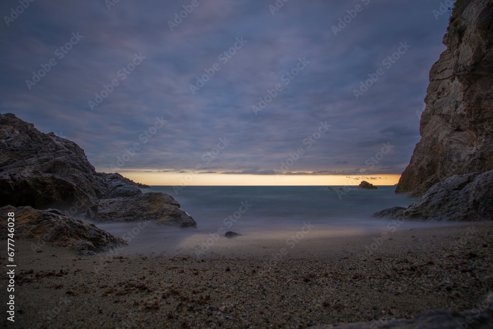 Punta Crena, bay in Liguria, shot without swimmers on a summer evening while the sun sets behind a veil of clouds. Sunset over the sea, long exposures. Varigotti, Liguria, Italy.