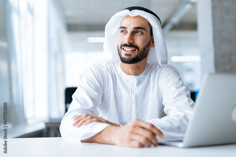 Handsome man with dish dasha working in his business office of Dubai. Portraits of a successful businessman in traditional emirates white dress. Concept about middle eastern cultures. Handsome man with dish dasha working in his business office of Dubai. Portraits of a successful businessman in traditional emirates white dress. Concept about middle eastern cultures.