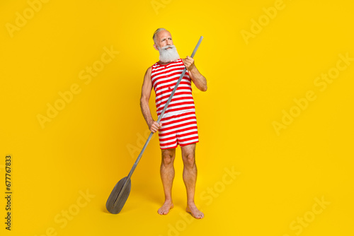 Full length photo of tourist old man kayaking looking beautiful natures surfing sap board with oar isolated on yellow color background photo