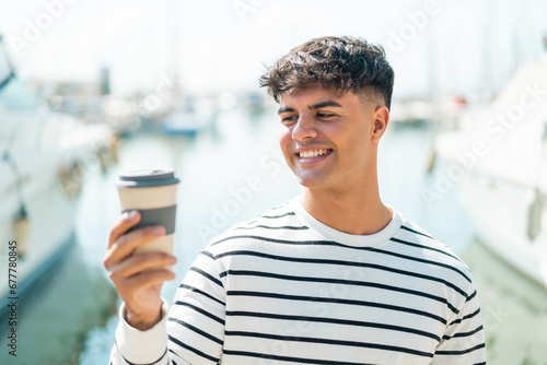 Young hispanic man holding a take away coffee at outdoors with happy expression