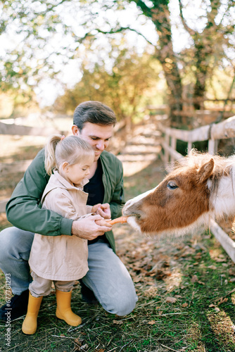 Smiling dad holding hand of little girl feeding pony through fence in park