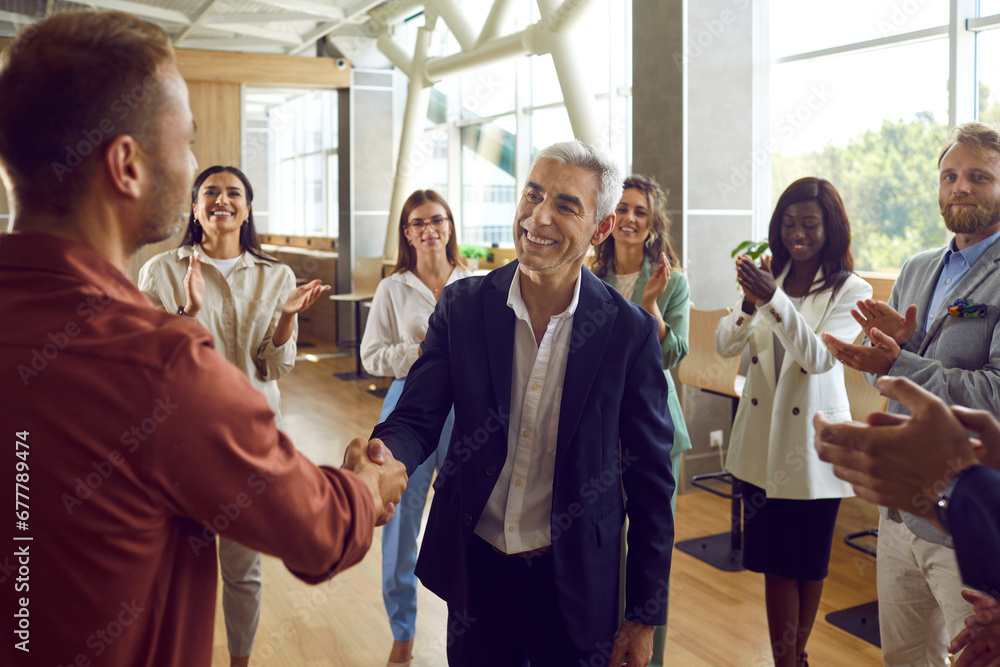 Two mature business man shaking hands celebrating success, making deal, business achievement, signing contract or greeting new employee with group of people applauding in the office. Two mature business man shaking hands celebrating success, making deal, business achievement, signing contract or greeting new employee with group of people applauding in the office.