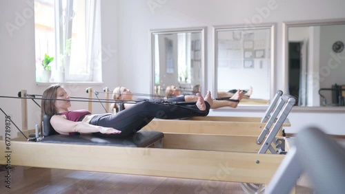 Two beautiful fit-shape females dressed sporty doing ABS muscles power exercises using pilates reformer machine in sport athletic gym. Active people training or medical rehabilitation concept image photo