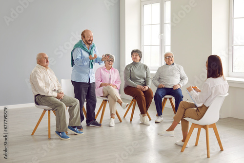 Senior people in group therapy. Happy retired old men and women sitting on chairs in a light office or retirement house and talking to a young friendly woman therapist with a clipboard
