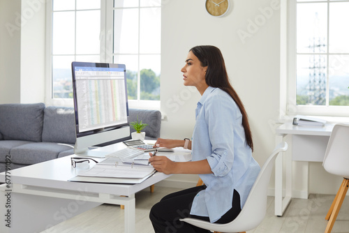 Portrait of a busy confident business woman accountant sitting at the desk and working on a pc computer with tables and charts analyzing company finances at office and looking at monitor screen. photo