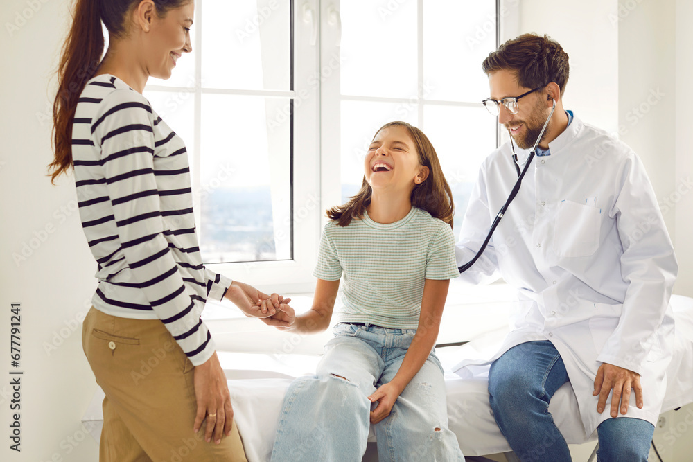 Obraz premium Male smiling family doctor with stethoscope examining child's lungs, breathing and heartbeat sitting on the couch with her mother in medical clinic. Pediatrician checking girl patient in exam room.