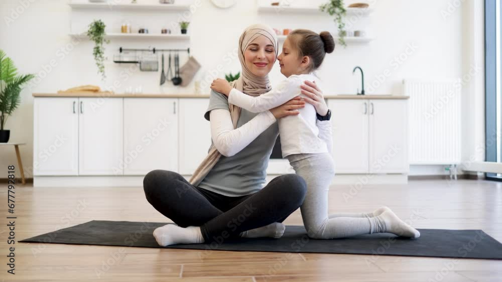 Adorable girl in sport outfit standing on knees on mat while trading positive energy with mom during exercises. Loving muslim female in hijab hugging daughter during fitness routine at home.