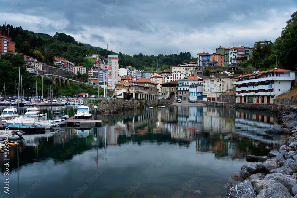 View of Mutriku village and its reflection in the harbor waters in the morning under a dramatic cloudy sky, Gipuzkoa, Basque Country, Spain