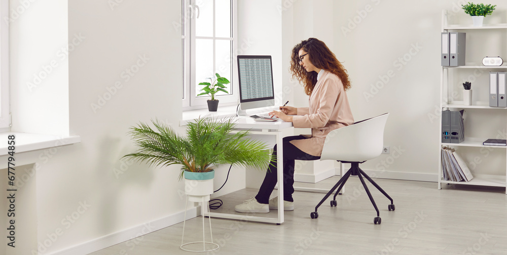 Side view portrait of a curly redhead young business woman accountant in glasses sitting at the desk and working on pc computer with tables and charts at the modern white office. Banner. Side view portrait of a curly redhead young business woman accountant in glasses sitting at the desk and working on pc computer with tables and charts at the modern white office. Banner.
