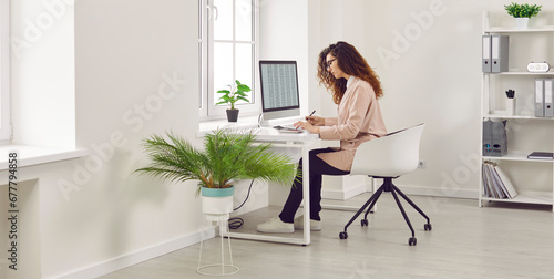 Side view portrait of a curly redhead young business woman accountant in glasses sitting at the desk and working on pc computer with tables and charts at the modern white office. Banner.
