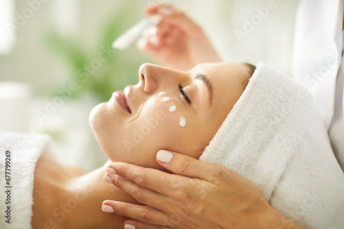 Close up beautician's hands applying anti-aging facial cream on woman client face to prevent wrinkles in spa salon. Skin care, cosmetic procedures for facial care and beauty treatment concept. photo