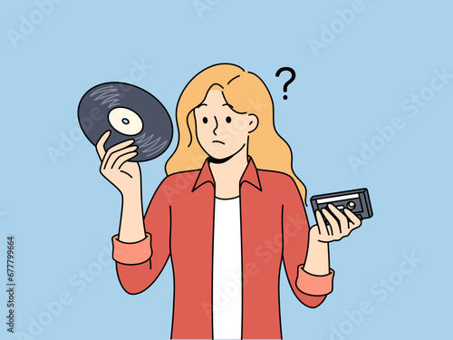 Girl with vinyl record and tape cassette looks confusedly at old-fashioned storage media with music. Woman feels confused and doesn know how to listen to music stored on vintage storage devices. photo