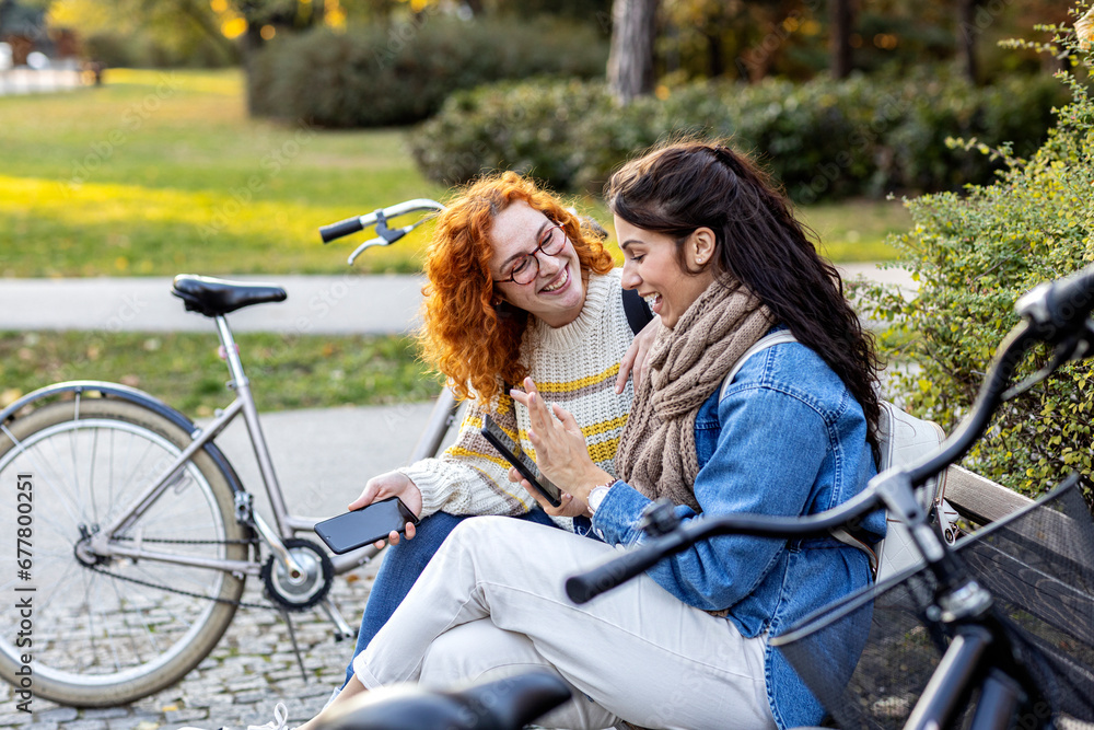 Two girl friends sitting on bench, looking at mobile phone and smile in public par with bicycle on background