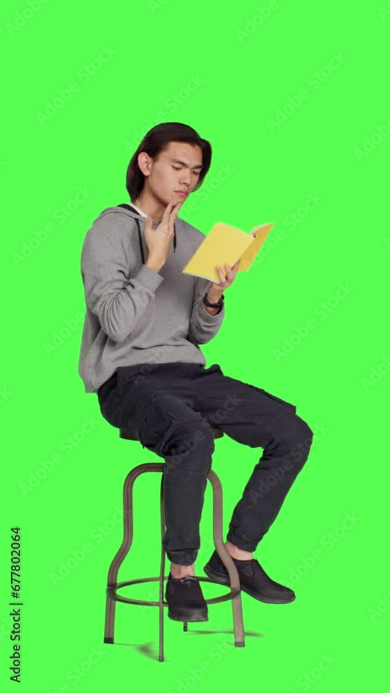 Side view of trendy person reading novel book on chair against full body greenscreen template, enjoying old school lecture hobby. Young asian adult holding literature or fiction book to read in studio Side view of trendy person reading novel book on chair against full body greenscreen template, enjoying old school lecture hobby. Young asian adult holding literature or fiction book to read in studio