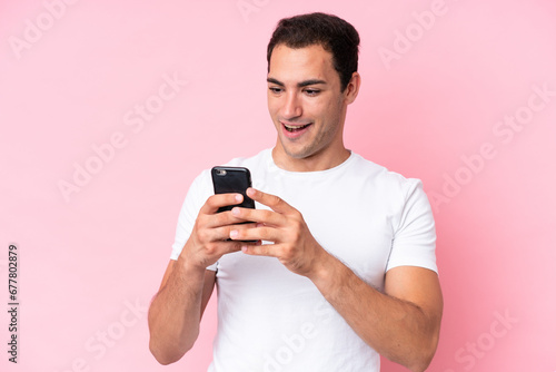 Young caucasian man isolated on pink background looking at the camera while using the mobile with surprised expression © luismolinero