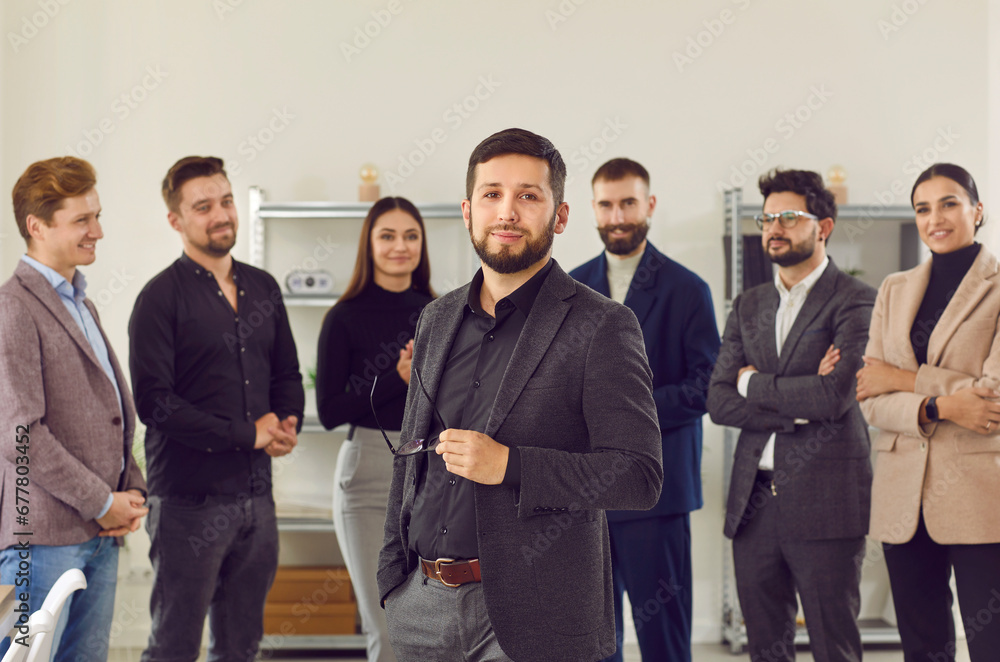 Smiling, happy businessmen stand around their colleague and greet new team member at work. Portrait of handsome, young man in business suit against the background of his business partners. Smiling, happy businessmen stand around their colleague and greet new team member at work. Portrait of handsome, young man in business suit against the background of his business partners.