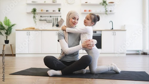 Adorable girl in sport outfit standing on knees on mat while trading positive energy with mom during exercises. Loving muslim female in hijab hugging daughter during fitness routine at home. photo