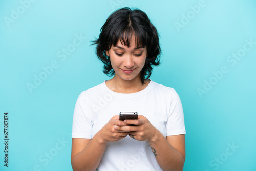 Young Argentinian woman isolated on blue background using mobile phone