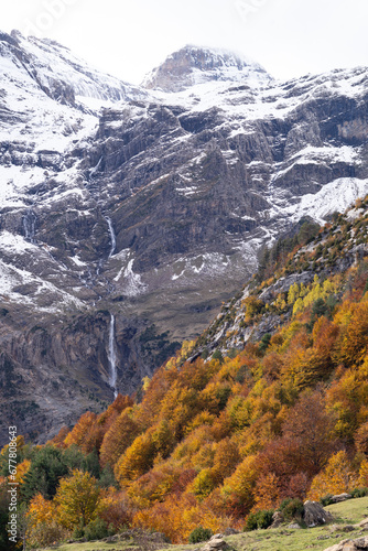 Waterfall in snow capped Massif of Monte Perdido with autumn color trees on the foreground.