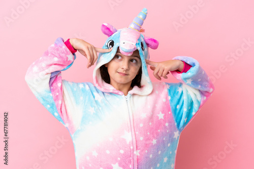 Little caucasian woman wearing a unicorn pajama isolated on pink background having doubts and thinking
