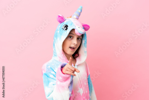 Little caucasian woman wearing a unicorn pajama isolated on pink background surprised and pointing front