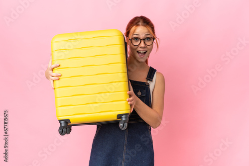 Teenager Russian girl isolated on pink background in vacation with travel suitcase and surprised photo