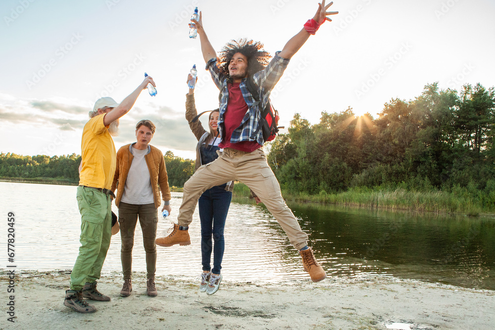 In this vibrant photo, a diverse group of millennial friends celebrates their arrival at a forest lake. Water bottles are joyfully raised in the air, capturing a moment of triumph. The excitement