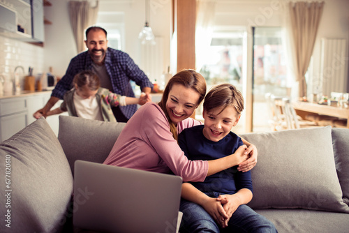 Family having fun at home together. Mother and son sitting on couch using laptop while father plays with daughter in background