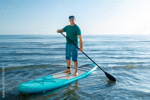 A middle-aged man in shorts and a T-shirt stands on a SUP board with a paddle near the sea.